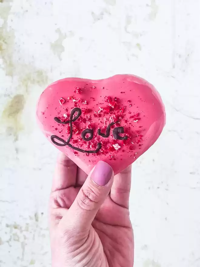 a hand holding a large pink heart shaped macaron with the word love written on it