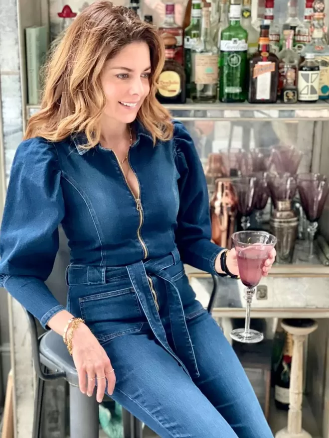 model wearing the bonnie denim blue jumpsuit sitting down by an open bar