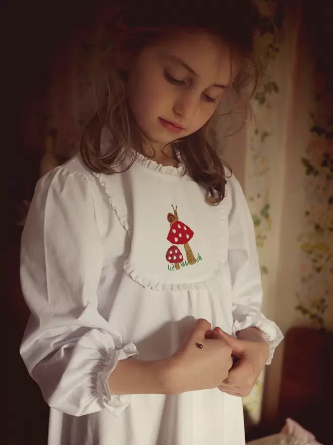 A girl holds a ladybird wearing a white nightie with embroidered toadstools