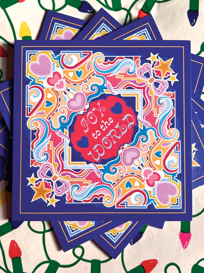 A square, blue Christmas card with an abstract, multi-coloured design including stars and hearts, with “Joy to the World” at the centre, is laid out on Christmas wrapping paper.
