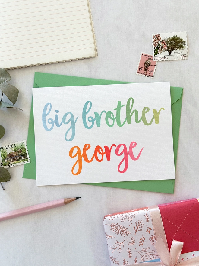 A personalised new baby sibling card with 'big brother' wording in pastel rainbow colours lays on a green envelope on a white desk.