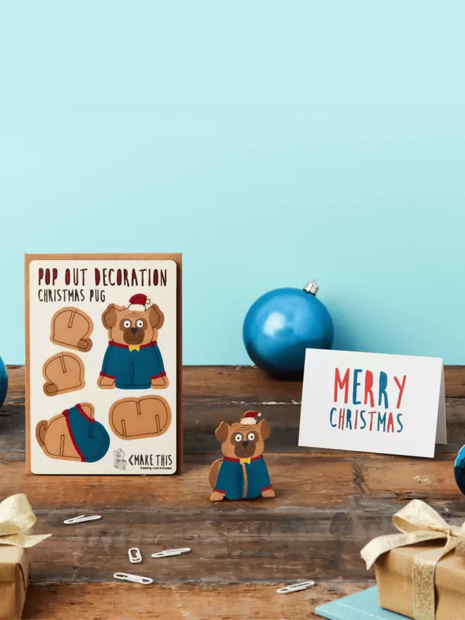 3D pug Christmas decoration and Merry Christmas text Christmas card and brown kraft envelope on top of a wooden desk in front of a light blue coloured background