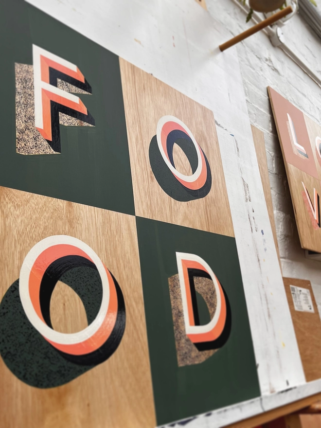 FOOD hand painted sign in coral, green and aubergine, against a white brick wall, in the artist's studio. 