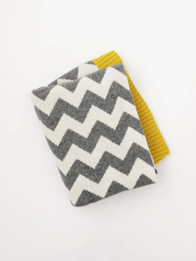  A folded junior blanket, photographed from above, showing a grey and white chevron pattern and a glimpse of mustard yellow trim.