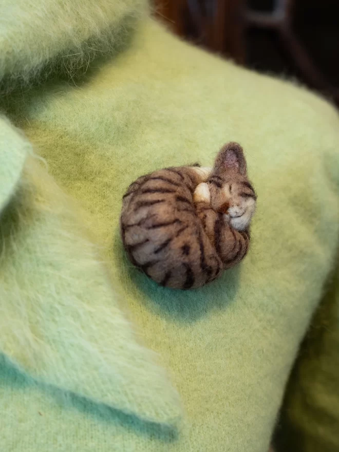 A needle-felted sleeping brown tabby cat brooch on a green fluffy jumper