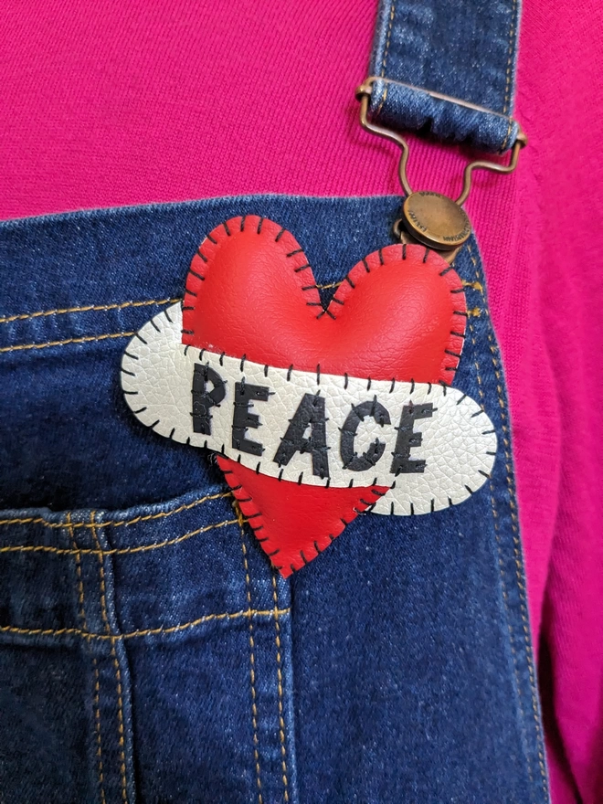 Hetty and Dave red leatherette heart brooch with PEACE in black lettering across a white scroll pinned onto denim dunagarees