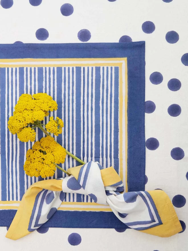 Navy stripe block printed tablemat with a navy spot napkin wrapped around two yellow flowers resting on top and over a navy spot tablecloth