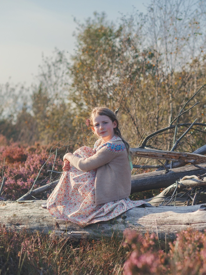 A girl in a beige cardigan with a fair isle yoke over a floral dress sits on a log surrounded by heather
