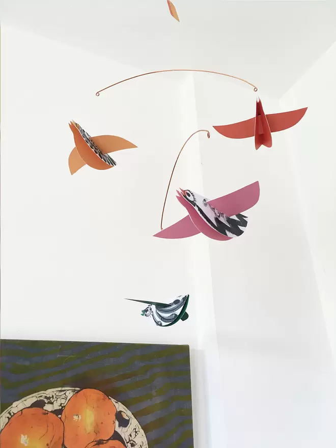 A mobile of four orange and pink paper birds flies above a half-seen canvas of oranges on a green background