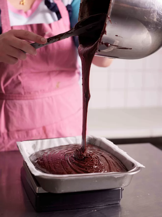 Founder Hetty in a pink apron pouring brownie mixture into a baking tray in the bakery