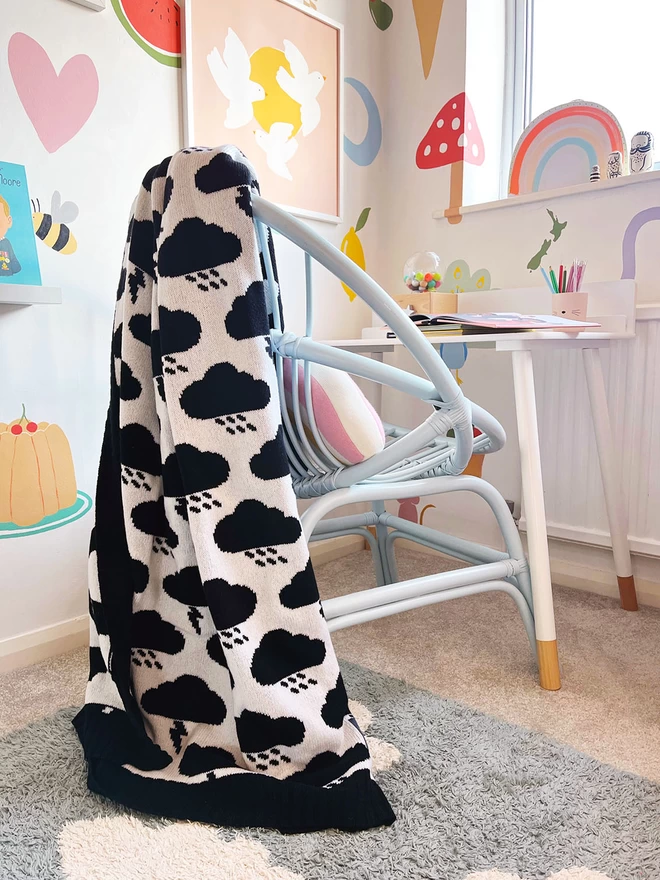 A corner of a bright and colourful childrens room showing a desk and chair. There are fun prints and murals painted on the walls. A black and white monochrome storm cloud blanket with rain clouds and lightning bolts on it is thrown over the back of the chair.