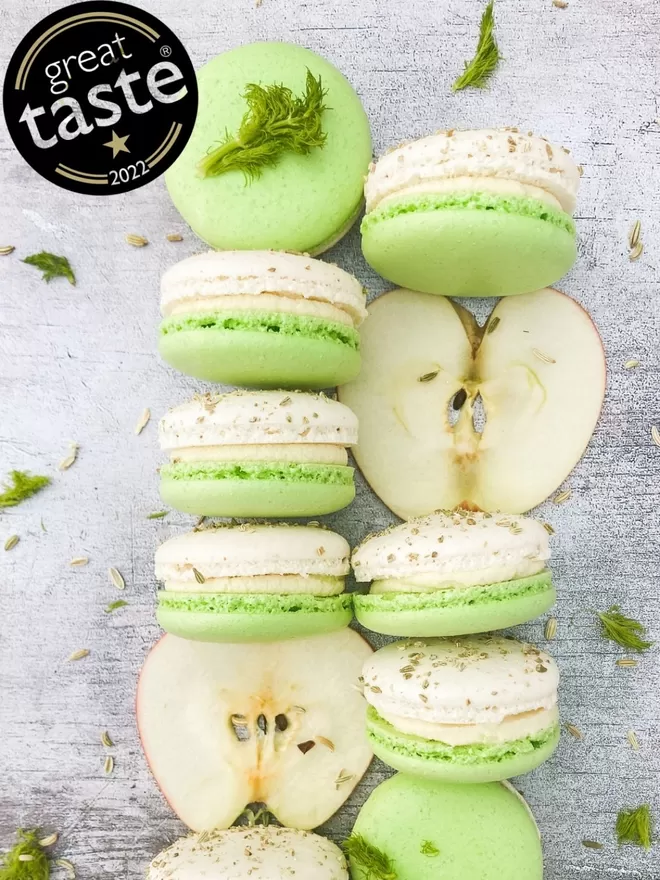 white & green macarons interspersed with thin apple slices on a grey background with fresh sprigs of fennel and fennel seeds. A great taste award logo in the top left hand corner