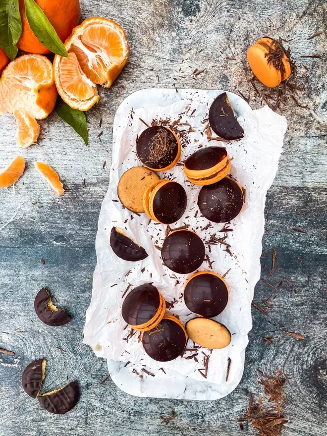 Orange Macarons with a shiny chocolate disc top in a pile with chocolate covered oranges on a wooden table with chocolate shavings