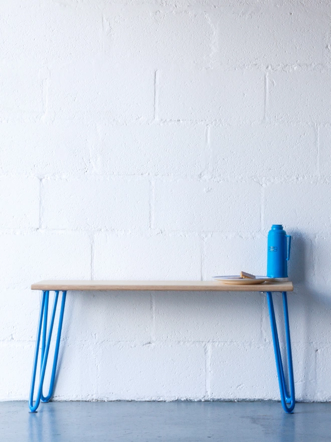 midcentury style hairpin leg bench with oak top and blue legs, against a white brick wall with a blue thermos flask sat on bench