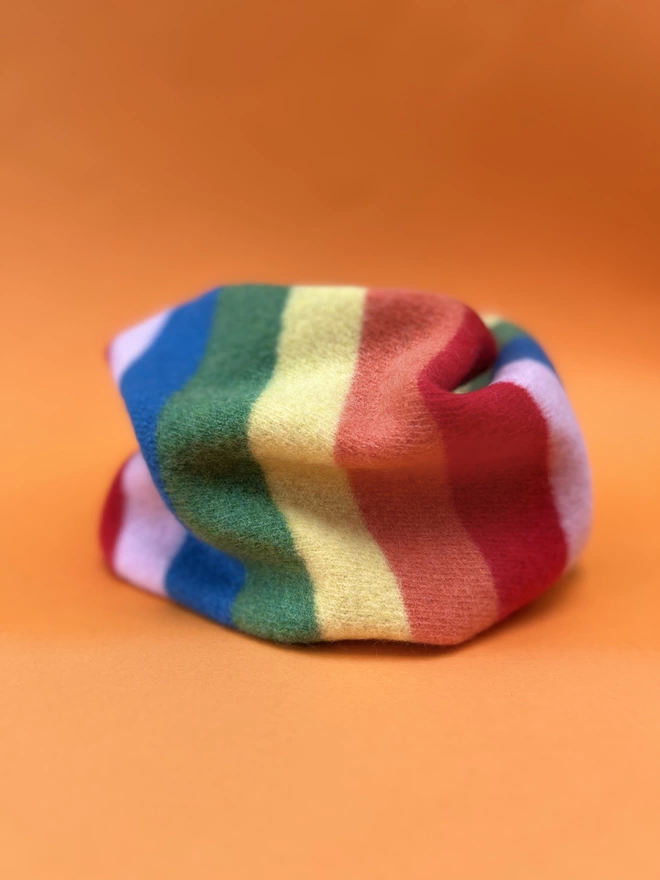 Rainbow knitted snood shown from the side on an orange background
