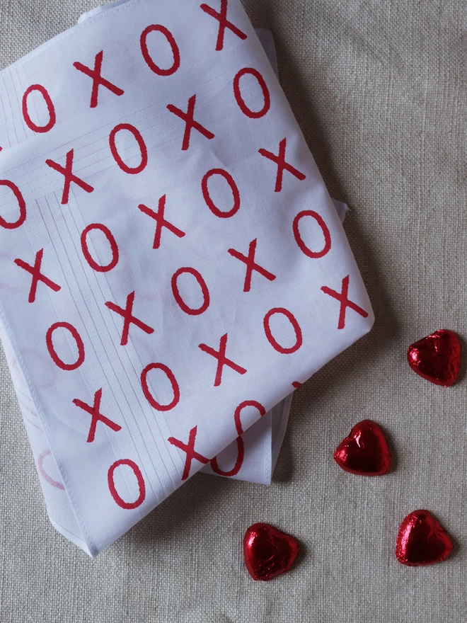 A Mr.PS Hugs and Kisses handkerchief printed in red laid on a linen tablecloth alongside some foil-wrapped chocolate hearts