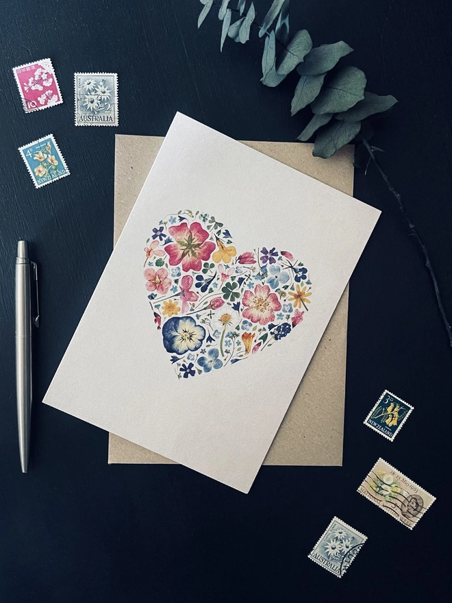 Floral Greetings Card with Pressed Flower Heart Design and Brown Kraft Envelope on Dark Charcoal Coloured Desk - Colourful Flower Postage Stamps, Silver Parker Pen, Dried Eucalyptus