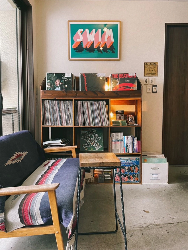 Art print of the word SWIM in 3d typography hangs above shelves of records with a sofa in the foreground at Chillout Coffee & Records in Tokyo Japan