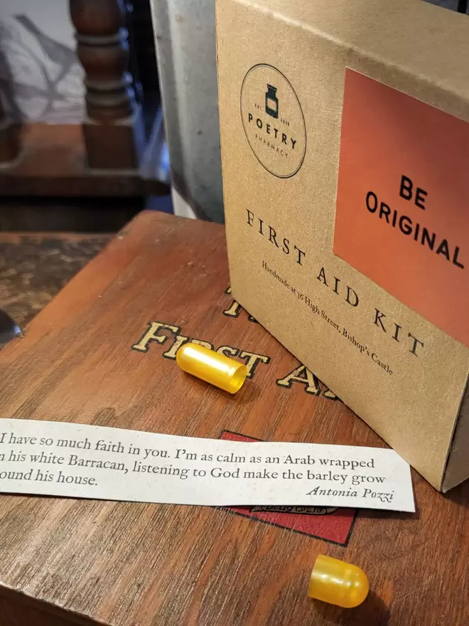 Cardboard Be Original First Aid Kit, closed, with empty golden pill capsule and a poem printed on banana paper. Displayed on vintage first aid kit