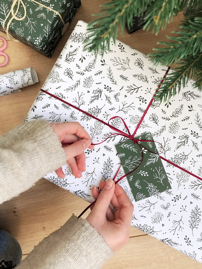 A gift wrapped in white wrapping paper with a Christmas botanical design lays on a wooden floor while a ribbon is being tied around it.