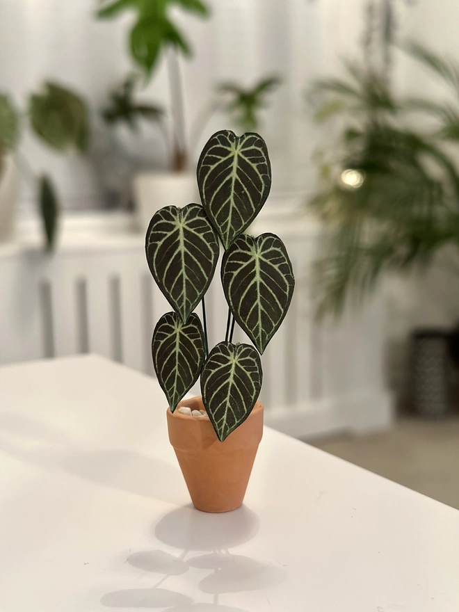 A miniature Anthurium Crystal Hope paper plant ornament sat on a white table with real plants in the background