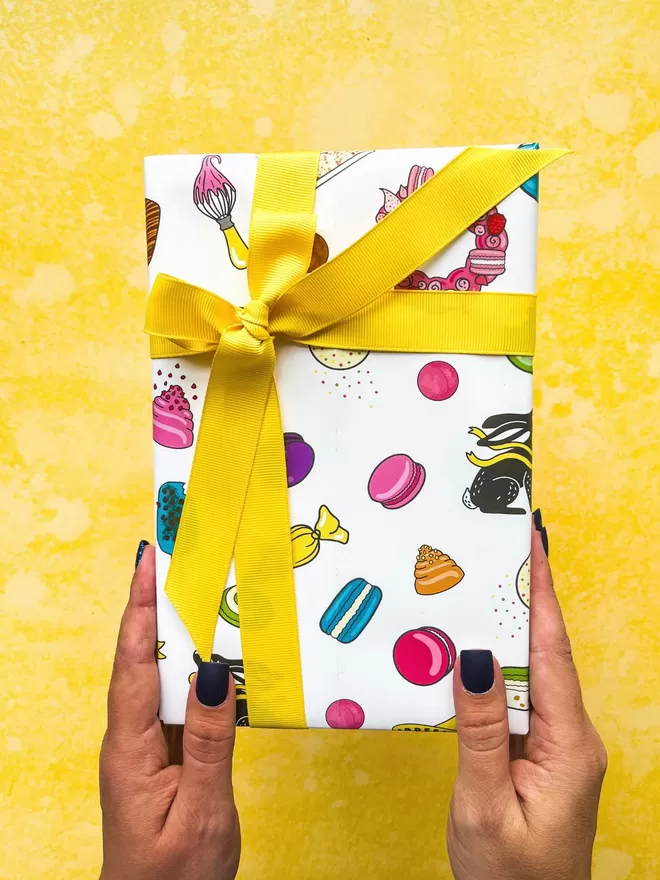 a female hand  with navy blue nails holding up a white gift box wrapped in a colourful macaron & chocolatey treats pattern, tied with a thick yellow ribbon  against a bright yellow background