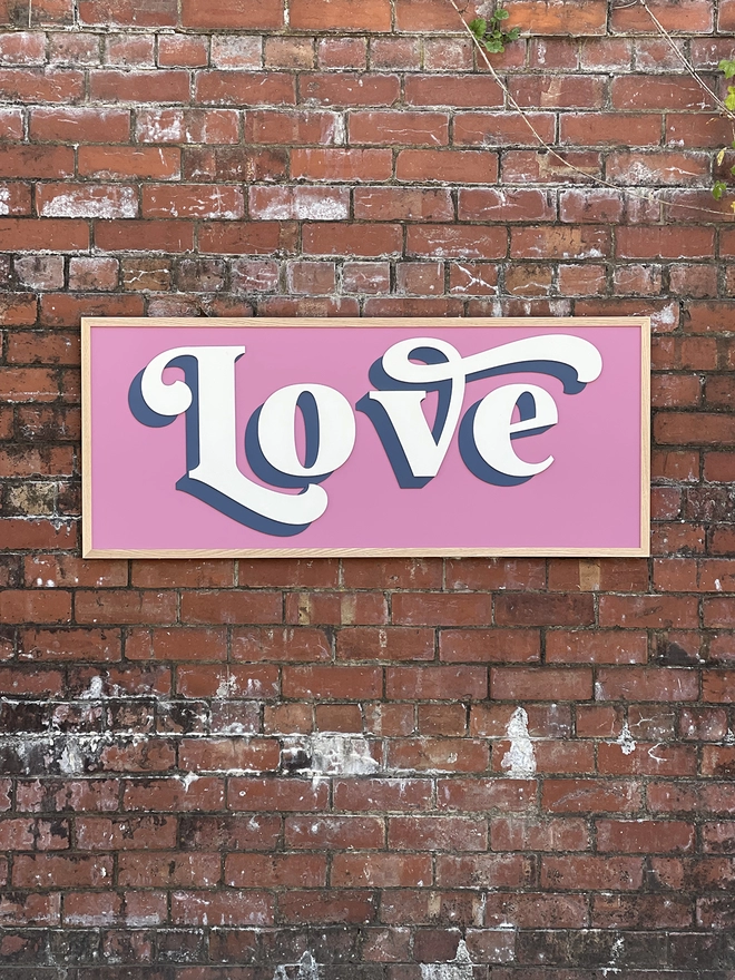 a painted wooden sign with a pink background hung on a brick wall which reads LOVE