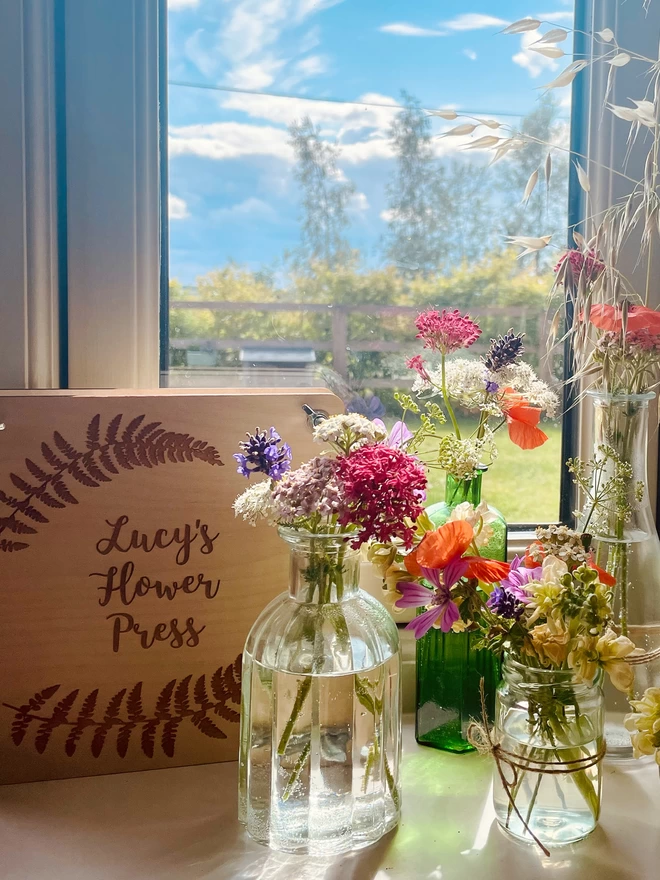 Windowsill with Wooden Flower Press Engraved with 'Lucy’s Flower Press' - Glass Bottles and Jars with Wildflowers - View of Sunny Garden with Blue Sky and Clouds Through Window