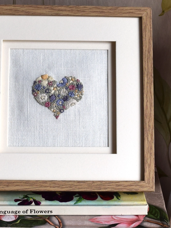 An embroidered Floral Meadow Heart, of Lavender Blues and Buttermilk yellow blossoms.  Displayed in an oak frame.