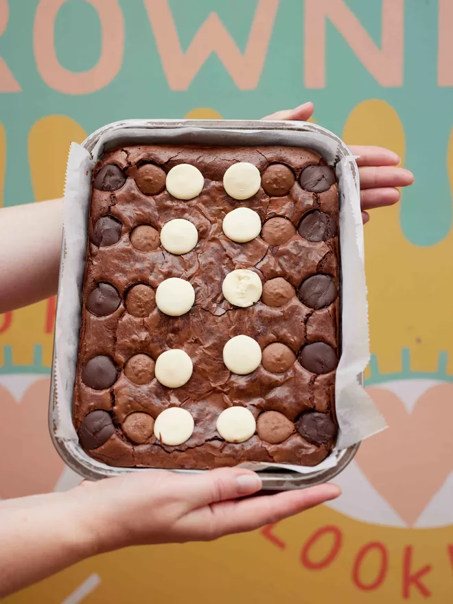 Freshly baked triple chocolate brownies in a baking tray being held in front of a colourful background