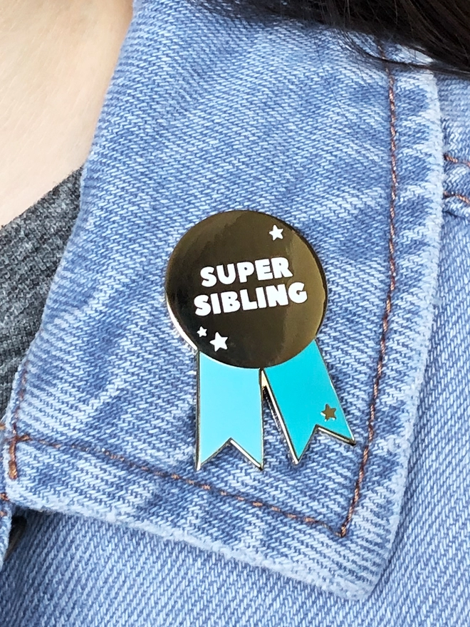 A turquoise and gold pin badge in the shape of a rosette is pinned to the collar of a denim jacket. It has the words "Super sibling" on.