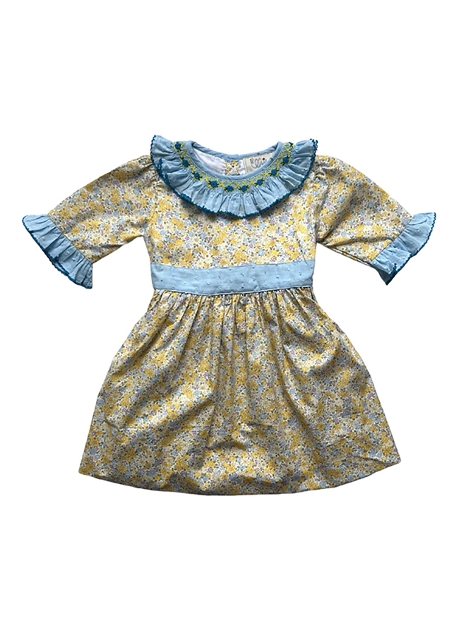 A yellow floral dress with a blue swiss dot frill collar and sleeves and a blue sash. Smocked detailing around the neckline.