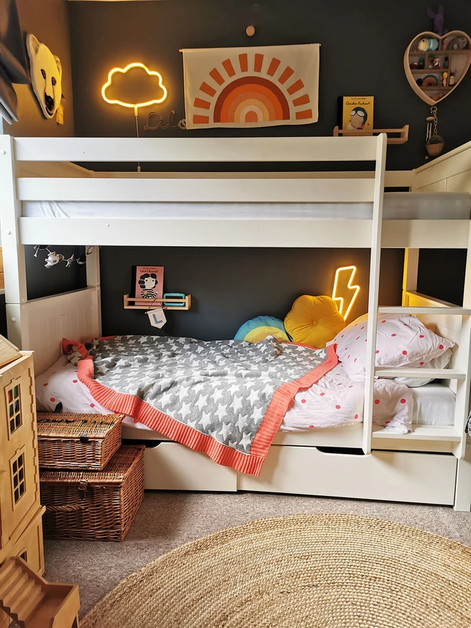 A view of white bunk beds in a childrens bedrrom. A grey knitted blanket with white stars on and a vibrant coral pink trim is draped over the bottom bunk. Above the beds hangs a fabric banner with an orange sunset, and cloud and lightning bolt neon night lights are turned on creating a warm and cosy feel. 