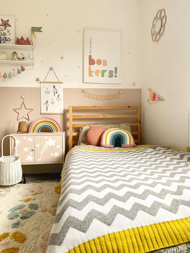 A view of a warm and vibrant childrens bedroom in muted rainbow colours showing a single bed with a grey and white chevron blanket with mustard yellow trim pulled up to the pillowcase.