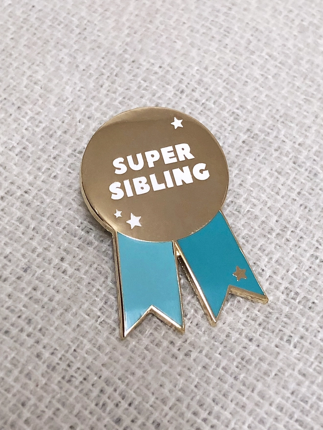 A turquoise and gold pin badge in the shape of a rosette is pinned to an ivory blanket. It has the words "Super sibling" on.