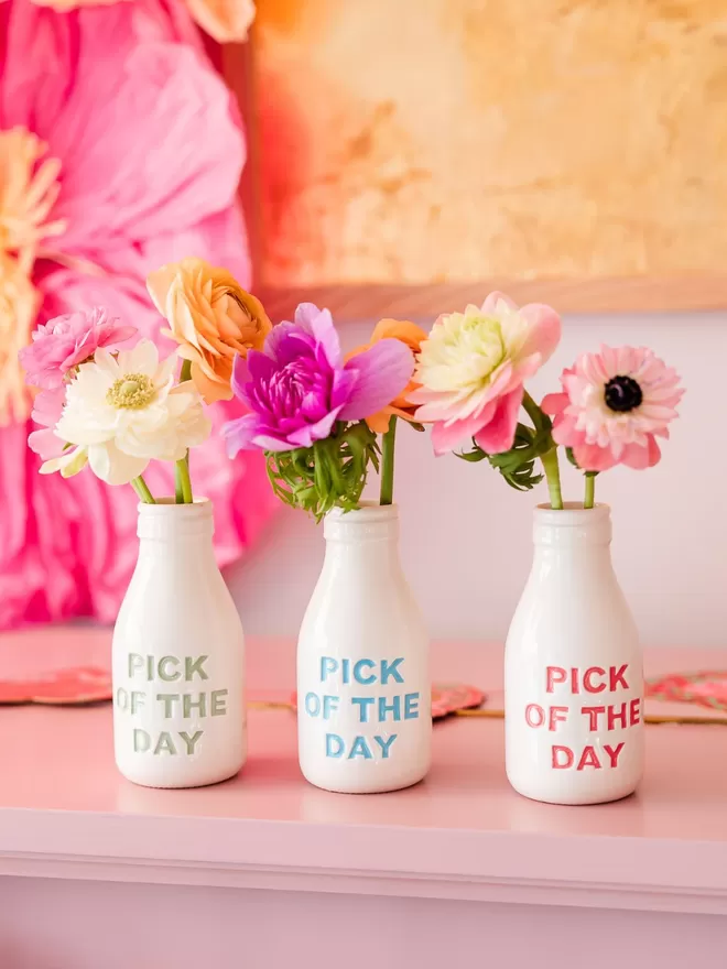 A handmade ceramic bottle/vase with ‘pick of the day’ lettering painted in blue, holding flowers.