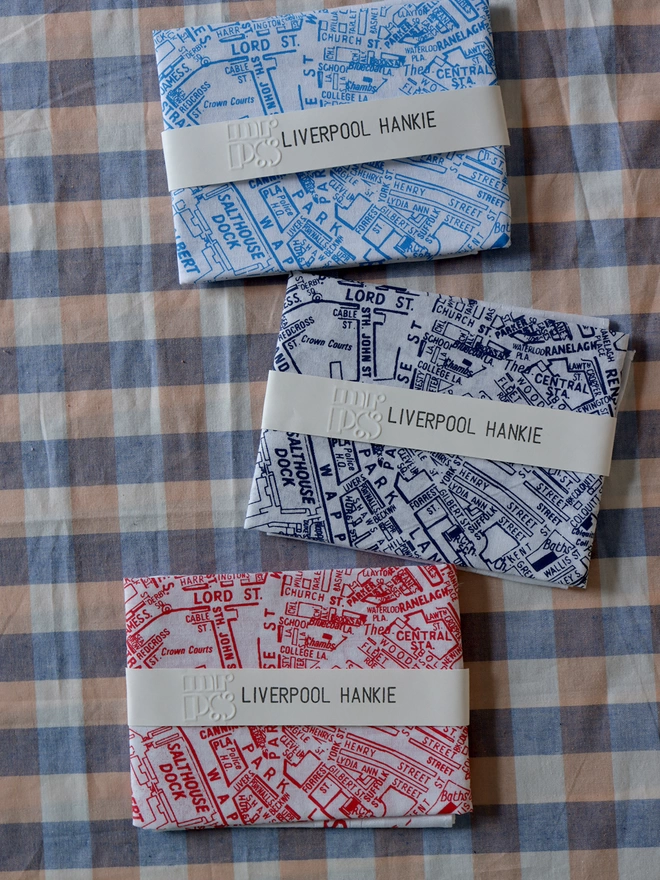 three folded Liverpool Hankies in sky blue, midnight blue, and red, showing the paper packaging labels,