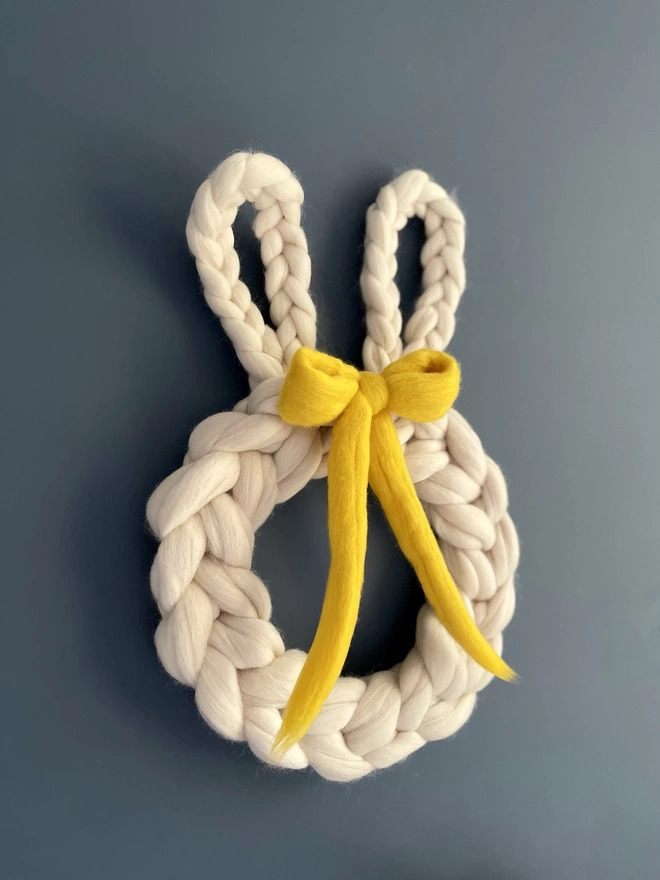 A light oyster cream woolly rabbit wreath is hung on a dark blue wall. It has a bright yellow bow. 