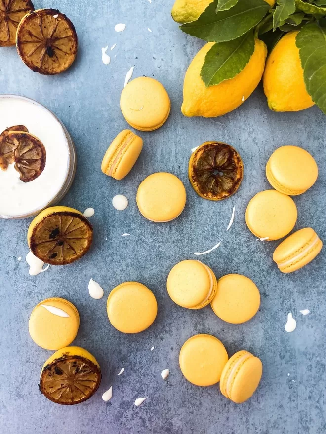 lemon yellow macarons on a blue surface with burnt lemon slices and white yoghurt splashes interspersed. With a pile of fresh lemons and lemon leaves in the top right corner
