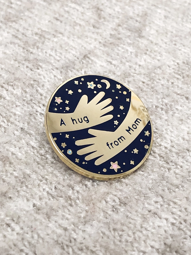 A navy blue and gold enamel pin badge with a hugging arms design and the words "A hug from Mom" is pinned to a beige blanket.