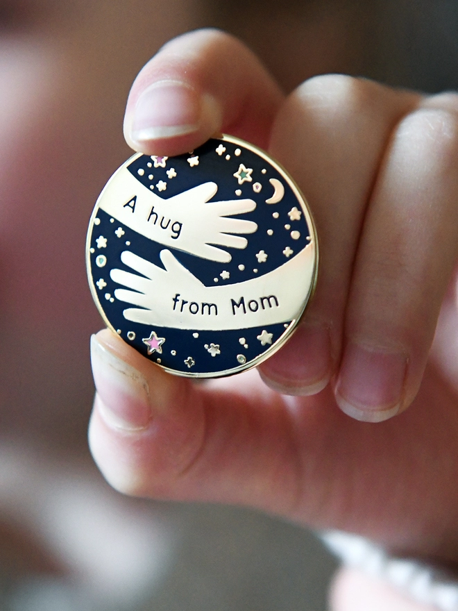 A navy blue and gold enamel pin badge with a hugging arms design and the words "A hug from Mom" is being held by in a small child's hand.