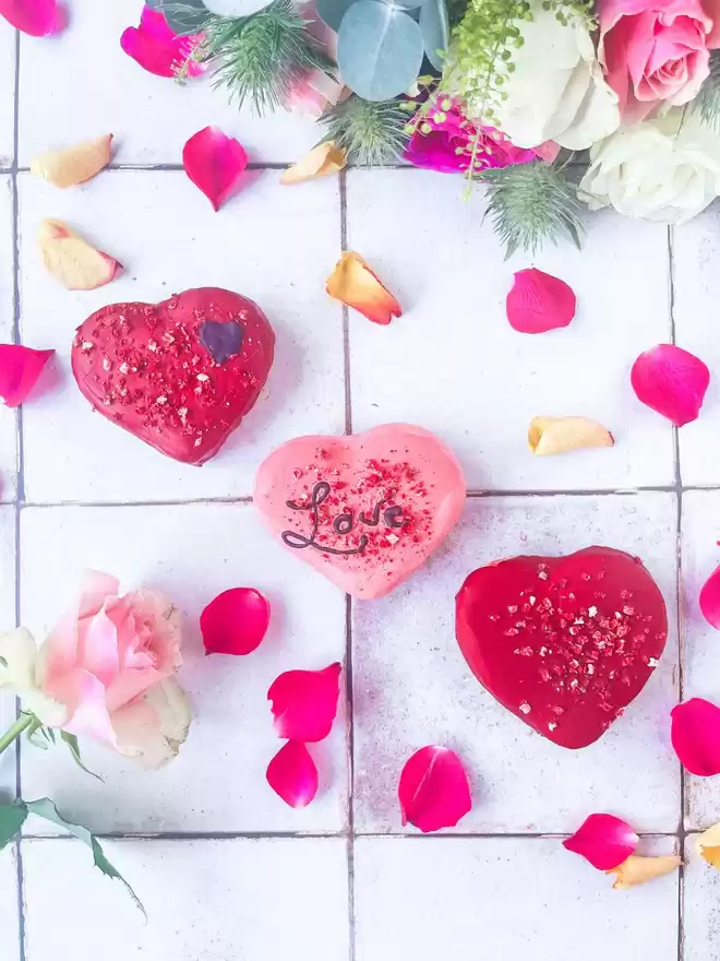 three large chocolate covered heart shaped macarons with a bouquet of flowers on a pink tiled background