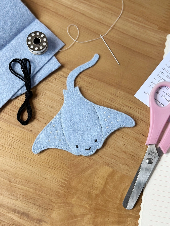 A soft blue felt manta ray has been sewn together and is on a wooden desk beside craft kit materials.