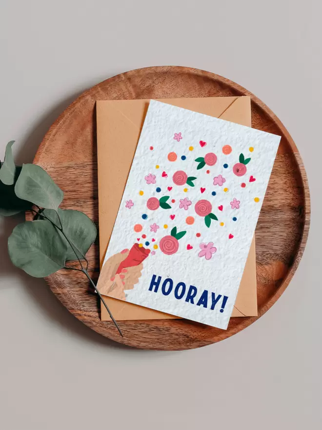Plantable Card with ‘Hooray’ in the bottom right of the card with an illustrated hand pulling a party popper with confetti and flowers coming out on a wooden tray next to a Eucalyptus branch