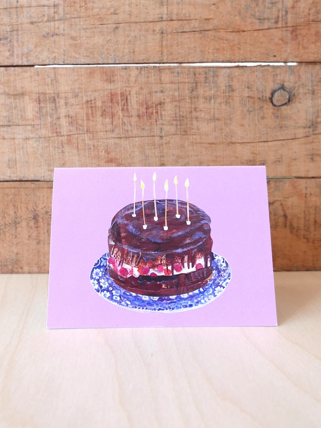 Landscape format birthday card of a chocolate cake & candles on a plate. It has a pink background. 