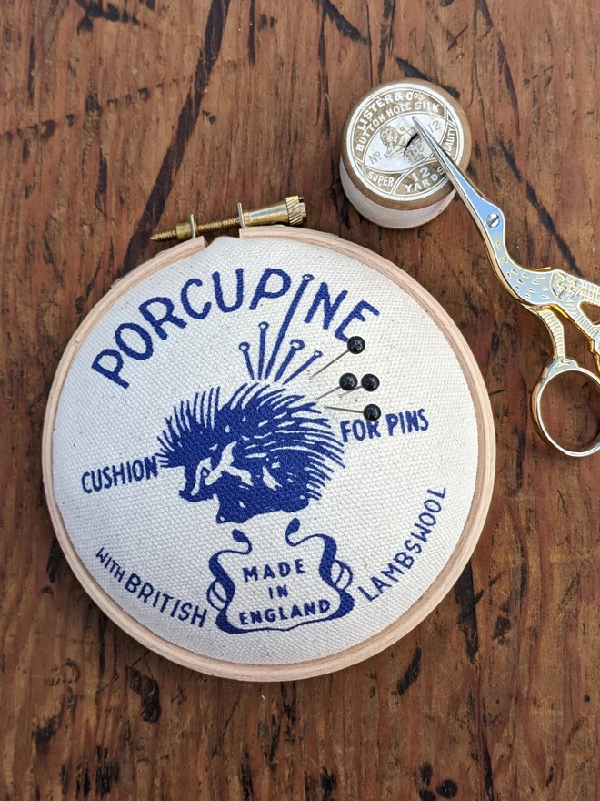 Royal blue porcupine embroidery hoop pin cushion shown with pins, scissors and cotton reel