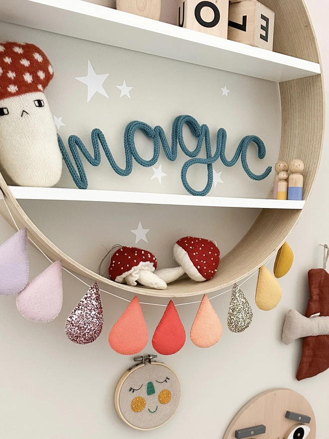 'magic' knitted wall word in 'petrol' blue on a shelf surrounded by kid's wall and shelf decorations. 
