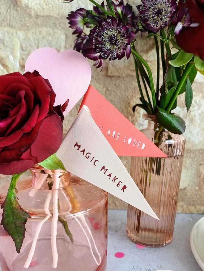 A closeup of two triangular flags displaying the papercut phrases "Magic Maker" and "Your Are Loved" The flags are placed in a bud vase with a red rose.
