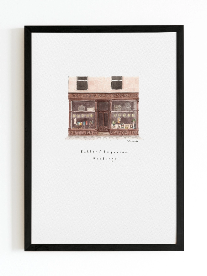 Watercolour illustration of Butler’s Emporium shop in Hastings, a brown shopfront with beautiful big windows full of treasures, the shop sells homewares and gifts carefully curated from small independent and local businesses. The painting is on a white piece of paper with the name written below. The illustration is a small watercolour painting in the centre of the white page and sits within a black frame. 