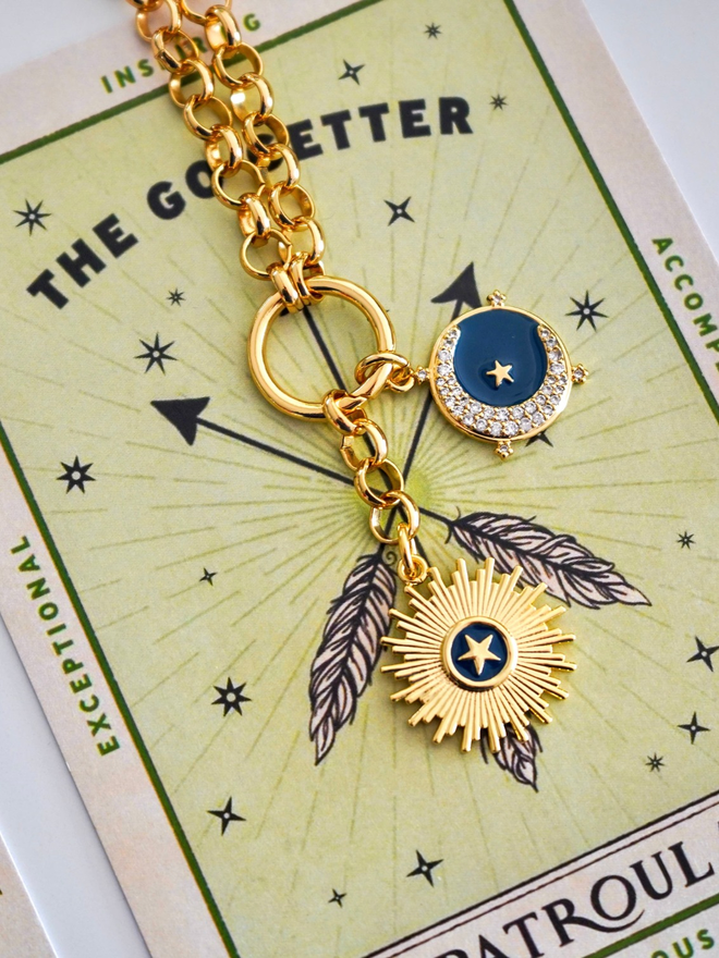 Gold belcher chain necklace with blue enamel star and moon charm laying on a green card with the words go getter